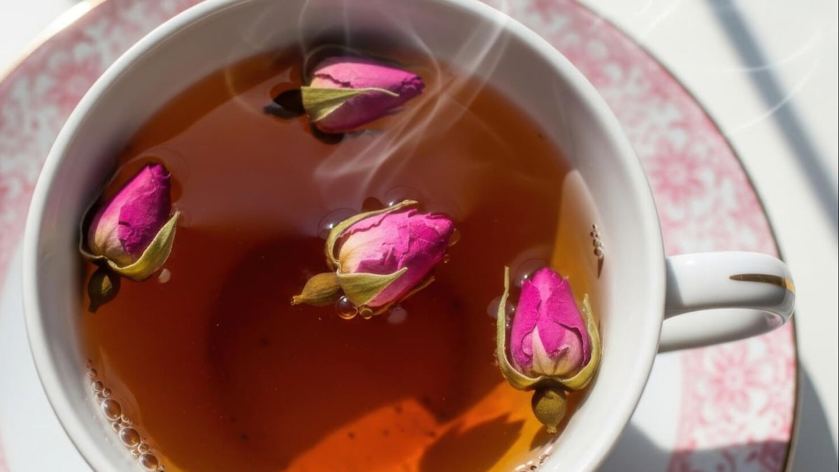 A white ceramic teacup filled with amber tea, floating pink rosebud tea blossoms, and visible steam, set on a matching pink floral saucer.